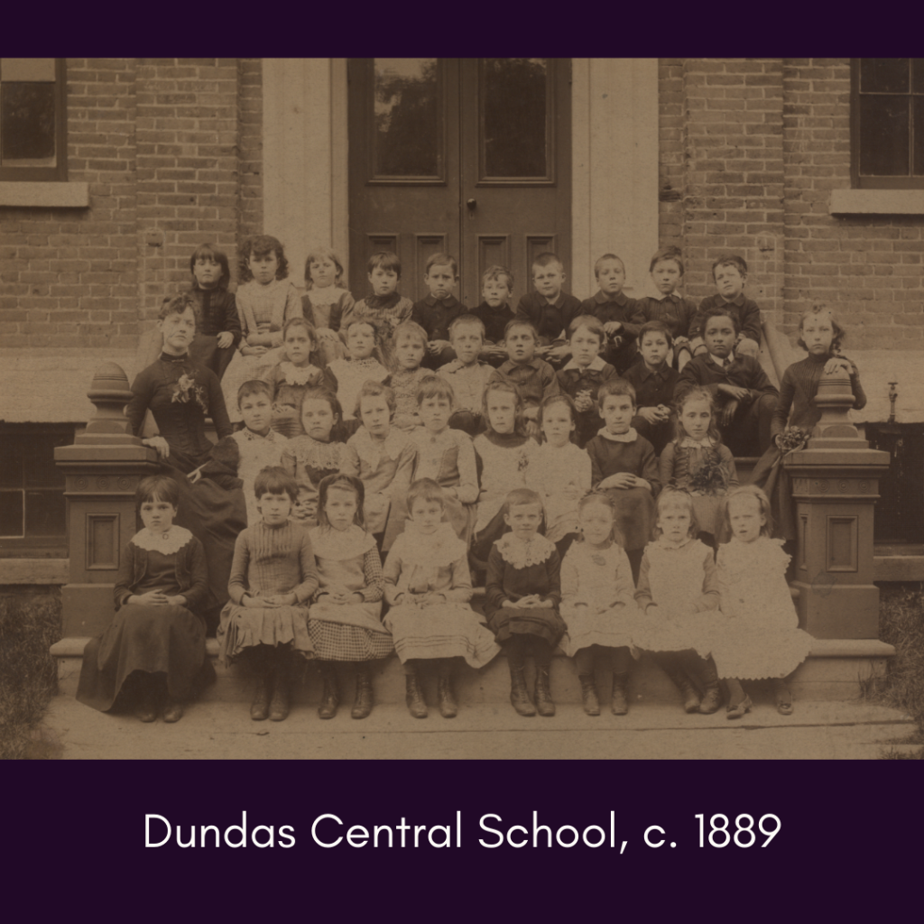 A sepia-toned class photo of girls & boys with neutral expressions sitting on the steps front steps of a brick building. 4 rows of students seated while their teacher leans against the railing, dressed in long skirts. The children’s clothes are period-appropriate, with lace-up boots, aprons & wide collars.
